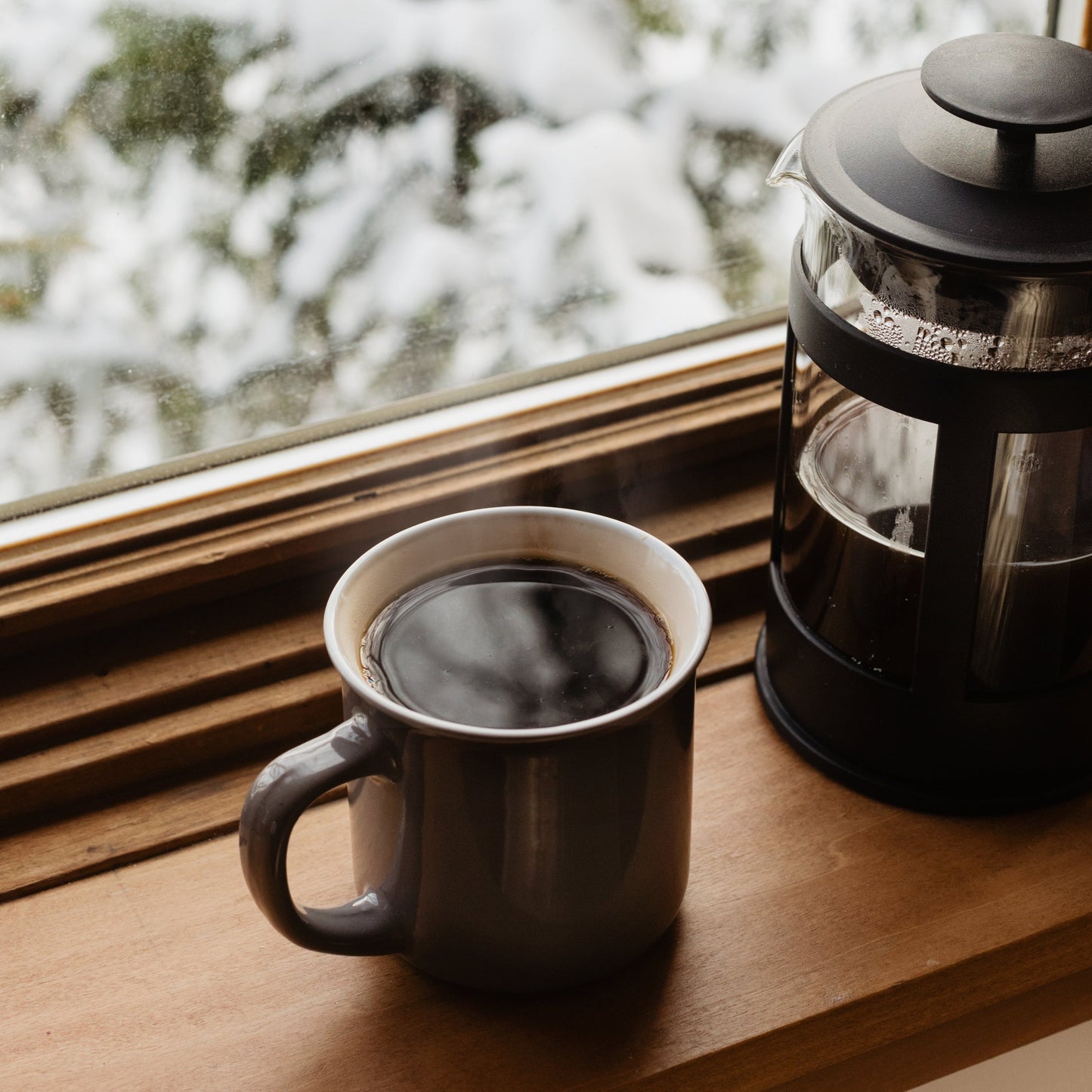 A fresh cup of coffee sits on a windowsill next to a french press coffeemaker. The window is blurring the snowy scene behind.