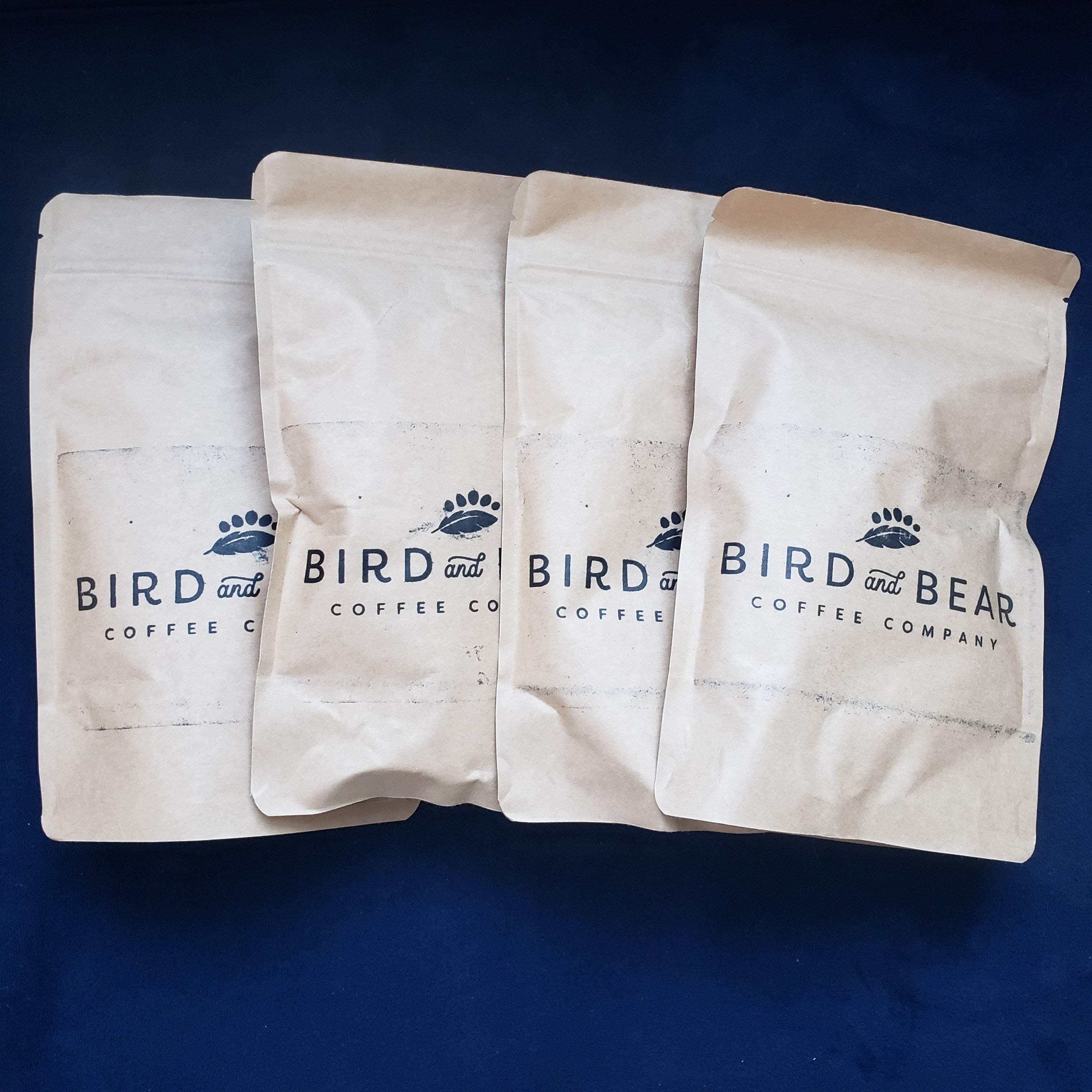 four sample size brown bags from Bird and Bear Coffee stacked on a plush navy blue background