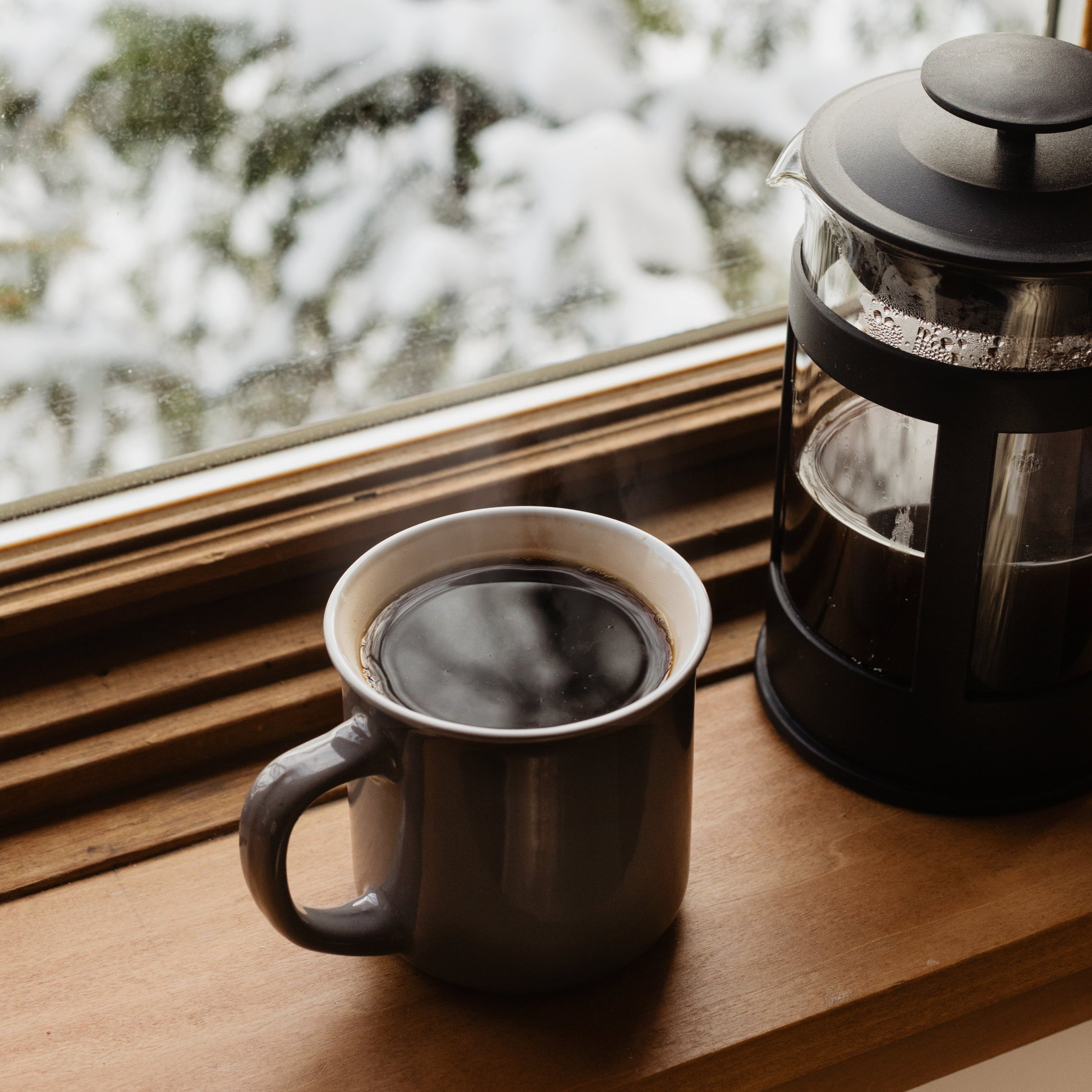 A fresh cup of coffee sits on a windowsill next to a french press coffeemaker.  The window is blurring the snowy scene behind.